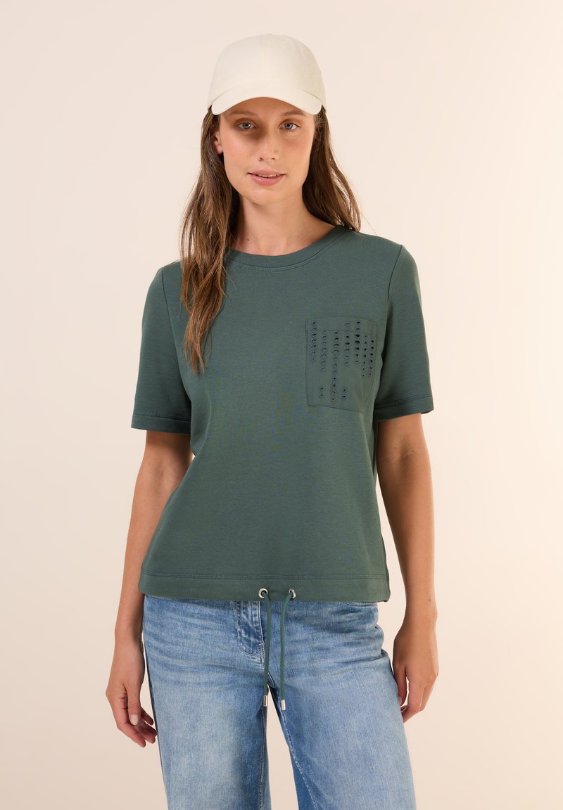 T-shirt vert olive à manches courtes, avec une poche texturée et un ourlet à cordon. Associé à un jean bleu clair.