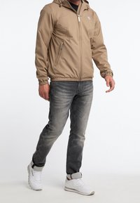 Béžová lehká bunda se zipem vpředu, nastavitelnou kapucí a elastickými manžetami, ve spojení s vybledlými šedými džíny a bílými teniskami.