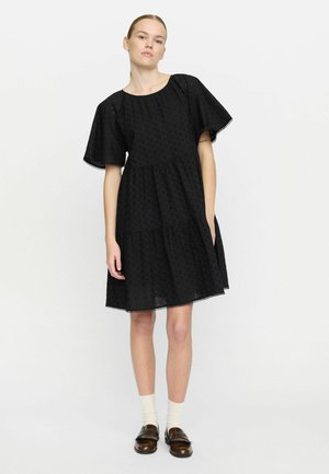 Schwarzes schichtetes Kleid mit kurzen, ausladenden Ärmeln, verziert mit bestickten Details und einem geschwungenen Saum. Getragen mit cremefarbenen Socken und braunen Loafers.