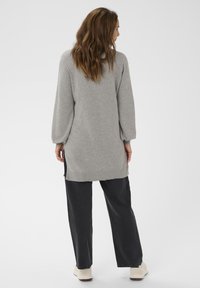 Žena s dlouhými hnědými vlasy, oblečená v šedých dlouhých pletených šatech, černých širokých kalhotách a bílých teniskách, stojící na bílém pozadí.