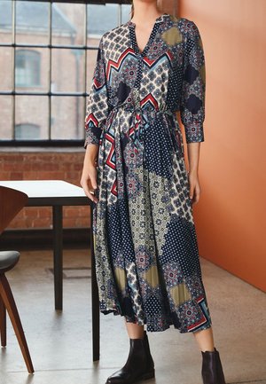 Mujer con vestido midi estampado de manga larga y botines negros, de pie en el interior cerca de una mesa y una ventana.