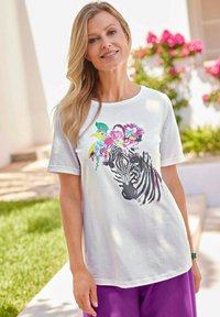 Witte T-shirt met korte mouwen, voorzien van een zwart zebra ontwerp versierd met kleurrijke bloemensaccenten op het hoofd. Draagt een paar paarse broek.