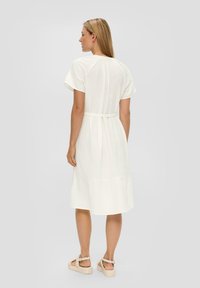 Witte, midi-lange jurk met korte mouwen, ronde halslijn, geraffineerde achterkant, riem in de taille en gelaagde zoom. Draagt met tan sandalen.