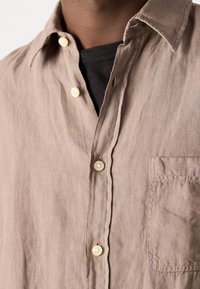 Jasnobrązowa lniana koszula z kołnierzykiem button-down, białymi guzikami i kieszenią na lewej piersi. Lekko pomarszczona tekstura.