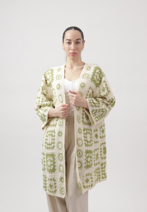 Mujer con un cárdigan largo y suelto de crochet color crema con patrones circulares y cuadrados verdes, camiseta blanca y pantalones beige, sobre un fondo liso.