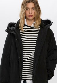 Čierna parka s umelou kožušinou na kapucni, skombinovaná s čierno-bielym pruhovaným sveterom pod bielou košeľou.