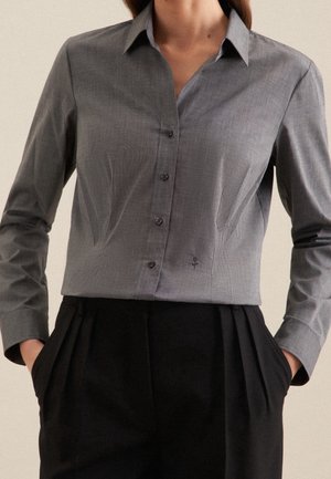 Vrouw draagt een nauwsluitend grijs overhemd met knopen, kraag en lange mouwen, gecombineerd met zwarte broek met hoge taille, handen in de zakken.