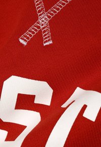 Tejido rojo con números impresos en blanco y un gran detalle de bordado blanco en forma de X. El textil presenta una textura con canalé.
