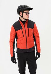 Rode en zwarte geïsoleerde jas met zakken aan de voorkant met rits, voorzien van een doorgestikt ontwerp en een hoge kraag. Draagt zwarte handschoenen en een helm.