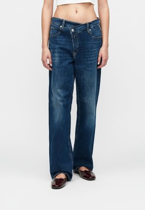 Kvinde iført løstsiddende mørkeblå jeans med åben lynlås i taljen og bordeaux flade sko, stående mod en ensfarvet baggrund.