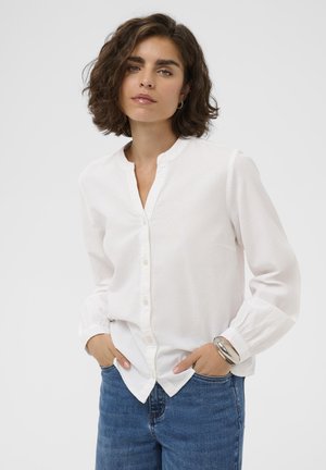 Biela dlhá košeľa s dlhými rukávmi a henley výstrihom, s gombíkmi vpredu a zhromaždenými manžetami, skombinovaná s modrými džínsami. Mäkko textúrovaná látka.