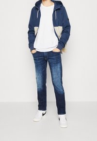 Navy blue hooded jacket with light beige accents, white t-shirt, dark denim jeans, and white sneakers with black logo.