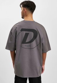 Grå oversized t-shirt med korte ærmer, der har et stort sort "D" logo og tekst på ryggen, lavet af blødt bomuldsstof.