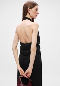 Μαύρο τοπ με λαιμόκοψη halter και ρέουσα λεπτομέρεια; το μοντέλο κρατά μια καημένη μπορντό τσάντα με υφή; λείο ύφασμα; μινιμαλιστικός σχεδιασμός; το μοντέλο γυρνάει背 από την κάμερα.