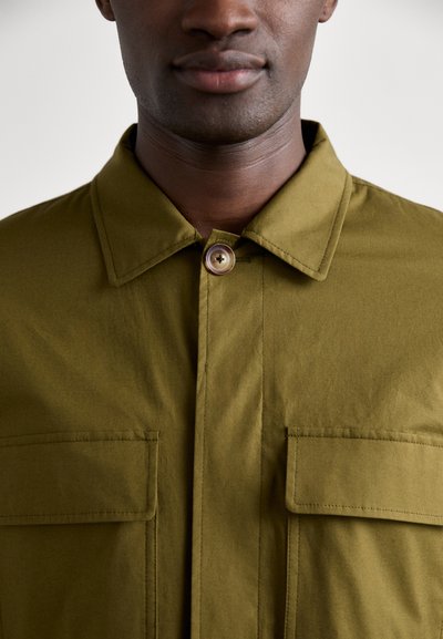 Oliven grøn, knap-op skjorte med spidse kraver og to frontlommer. Glat tekstur med synlige sømme og en enkelt rund knapdetalje.