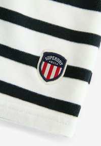 Randig tyg i vitt och mörk marinblått, med en cirkulär emblem i rött, vitt och blått som visar "SUPERDRY" och "N.Y.C."