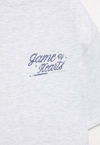 T-shirt gris à manches courtes en tissu doux, avec une broderie bleu marine portant l'inscription "Game of Hearts" accompagnée d'étoiles décoratives.