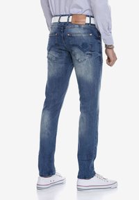 Jeans aus Denim mit einem verblassten blauen Finish, gerade Passform, zwei Gesäßtaschen mit dekorativer Stickerei, getragen mit einem weißen Gürtel und weißen Turnschuhen.