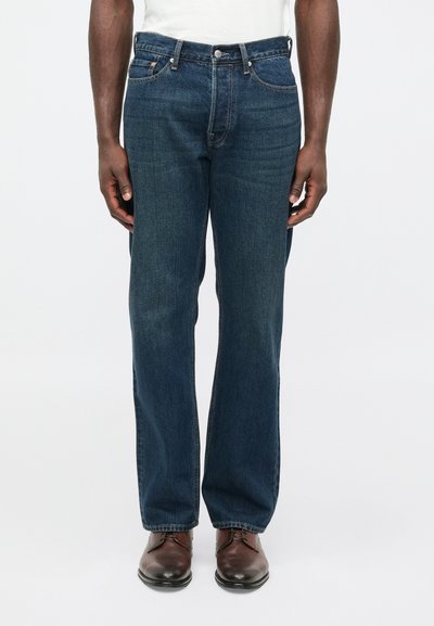 Donkere blauwe spijkerbroek met een rechte pijp, vijfzakkenmodel en contrasterende stiksels. Gecombineerd met bruine leren schoenen.