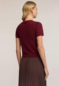 Burgundský svetr s krátkým rukávem a přiléhavým střihem, spárovaný s hnědou plisovanou sukní. Svetr má jemnou strukturu a žebrovaný lem.