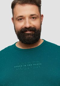Türkise T-Shirt mit Rundhalsausschnitt, mit einem strukturierten Druck, der "S.OLIVER SPACE IS THE PLACE" in passender Farbe zeigt. Gesprenkelte Texturdetails.