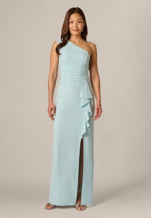 Kobieta w jasnoniebieskiej, asymetrycznej sukni wieczorowej z falbaną i rozcięciem z przodu, stoi na jednolitym tle.