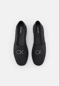 Sorte slip-on sko med vævet tekstur, der har et fremtrædende sølvfarvet CK-logo og en flad sål. Enkel design uden yderligere pynt.