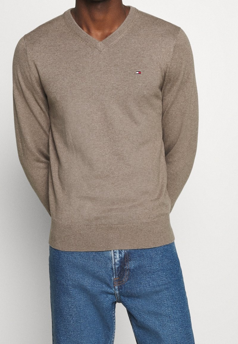 Homme portant un pull beige à col en V avec un petit logo sur la poitrine et un jean bleu, debout devant un fond clair uni.