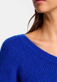 Maglione blu a maglia con scollatura ampia, caratterizzato da un motivo testurizzato e orlo a coste. Il tessuto appare morbido e caldo.