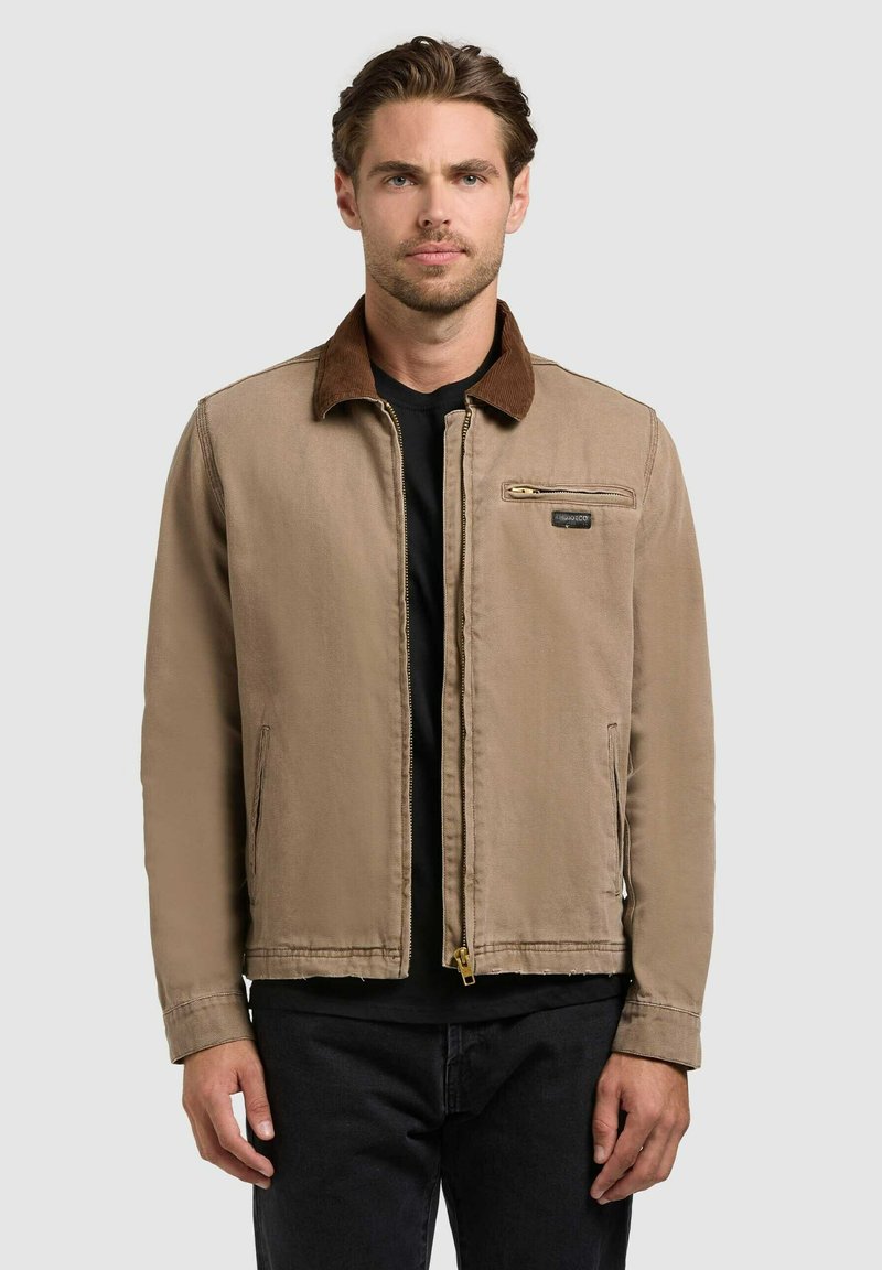 Homme portant une veste beige à fermeture éclair avec un col marron et une poche zippée sur la poitrine, associée à une chemise noire et un pantalon noir.