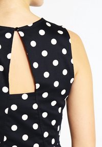 Černé šaty s bílými puntíky, bez rukávů, s otvorem ve tvaru klíčové dírky na zádech, vyrobené z hladké tkaniny.
