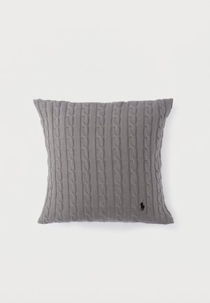 Coussin en tricot gris avec motifs en câbles verticaux et un petit logo noir brodé près du coin inférieur droit sur un fond blanc.