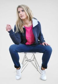 Marinefarbener Zip-Hoodie mit einer grauen gefütterten Kapuze, kombiniert mit einem pinken T-Shirt mit Strassdesign, dunkelblauen Jeans und weißen Sneakern.