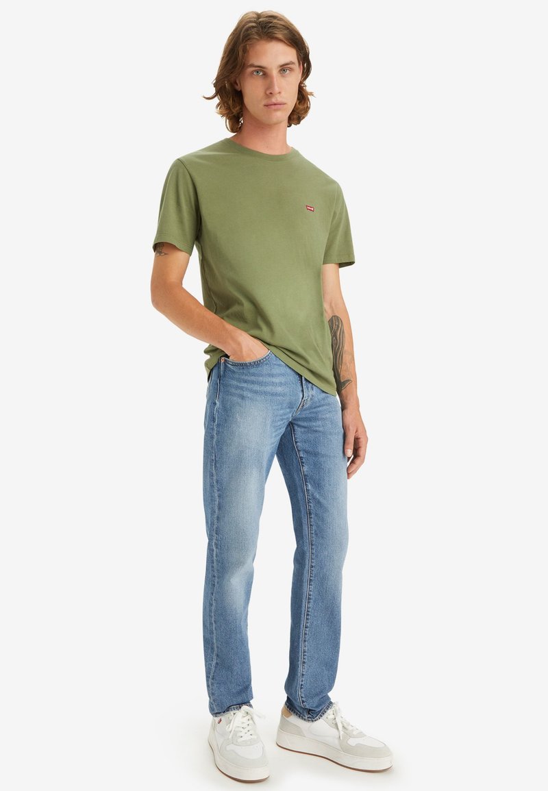 Camiseta verde de manga corta con un pequeño logo rojo, combinada con vaqueros azul claro y zapatillas blancas. Diseño sencillo, ajuste casual.