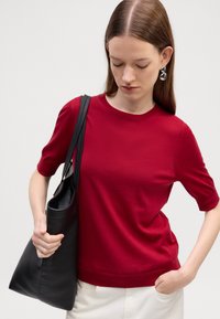Červený svetr s krátkým rukávem a kulatým výstřihem, měkká textura, v kombinaci s černou koženou taškou. Ruce modelky jsou umístěny na tašce a kapse.