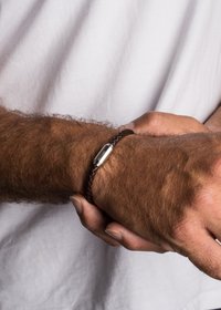 Braccialetto in pelle intrecciata marrone con chiusura metallica e dettagli incisi, indossato su un polso con peli chiari.
