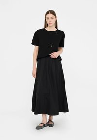 T-shirt nera in cotone con maniche corte e dettaglio con coulisse, abbinata a una gonna nera fluente. La modella indossa ballerine nere semplici.