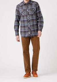 Man som bär en knappad blå och brun rutad skjorta, bruna byxor och bruna läderskor med snörning, stående mot en vit bakgrund.