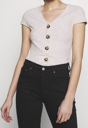 Vrouw draagt een lichtroze geribd topje met korte mouwen, vier grote bruine knopen en een zwarte broek met hoge taille.