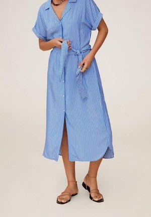 Person bindet Gürtel eines blau-weiß gestreiften, knielangen Hemdkleides mit vertikalen Streifen, trägt beigefarbene, flache Sandalen mit Riemchen und eine Halskette.