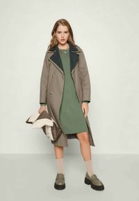 Trench coat longo, castanho claro, com colarinho escuro, usado sobre um vestido verde. Sapatos de plataforma pretos e meias são visíveis. Textura suave, detalhes mínimos.