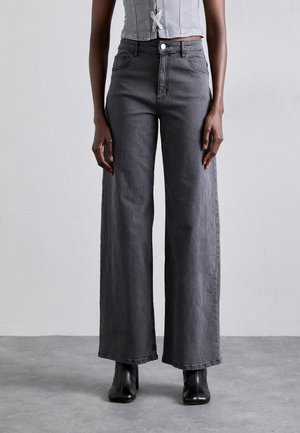 Personne portant un jean gris foncé taille haute à jambes larges, des bottines noires et un top court gris clair, debout devant un fond uni.