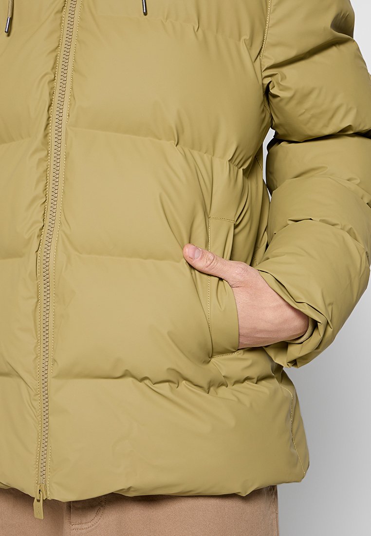 Persoon in een beige donsjack met een hand in de voorzak, rits dicht, zichtbare stiknaden van de jas en gevoerde mouwen.