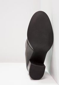 Černá kožená bota s hladkým svrškem a strukturovanou gumovou podrážkou. Podpatky jsou zúžené a široké a mají jemně vroubkované detaily na spodní části.