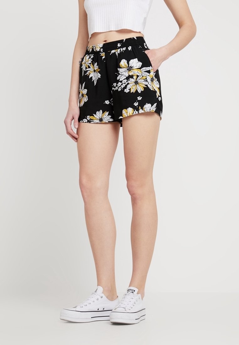 Femme portant un short noir à motifs floraux, un top blanc court sans manches, et des baskets blanches à plateforme, debout avec une main dans la poche.