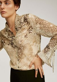 Camisa beige de botones con un patrón de estampado animal marrón, mangas largas con puños acampanados y tejido ligero y transparente. La prenda cuenta con un lazo decorativo en los puños.