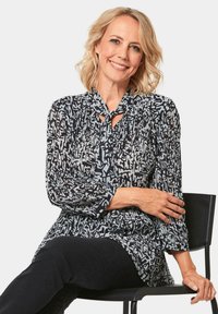 Zwart-witte blouse met een patroon, een striknek, geplisseerde textuur en lange mouwen. Draag over zwarte broek terwijl gezeten.