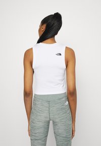 Cropped white tank top with a round neckline, featuring a small black logo on the back. Paired with textured green leggings.