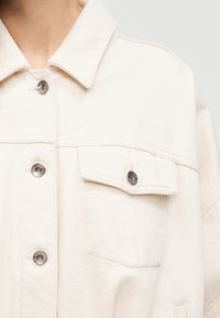 Jeansjacke in Cremefarben mit spitzem Kragen, zwei Brusttaschen und silbernen Druckknöpfen. Verfügt über eine strukturierte Stoffoberfläche.