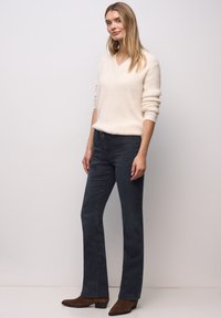 Pull à col en V couleur crème en matière douce, associé à un jean évasé en denim foncé avec poches avant et fermeture à boutons. Bottines marron.